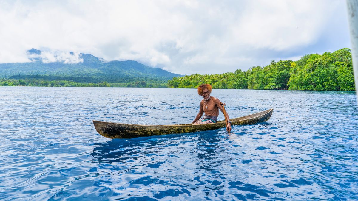 An elderly man paddles a wooden canoe in calm blue waters, surrounded by lush green trees and mountains under a cloudy sky.