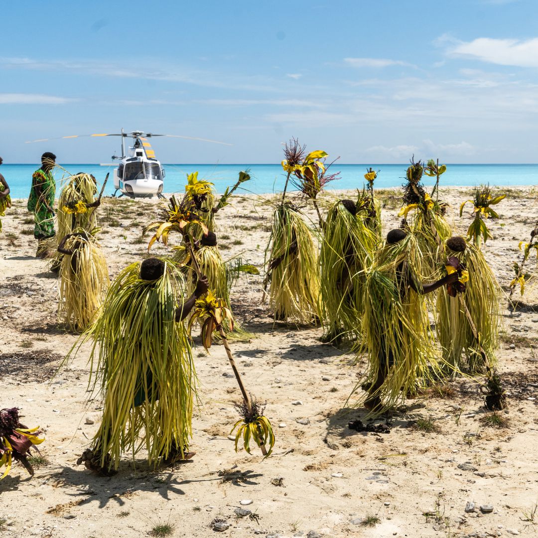 A group of people dressed in traditional attire made from palm fronds performs a dance on a sandy beach with a helicopter in the background and a clear blue ocean.