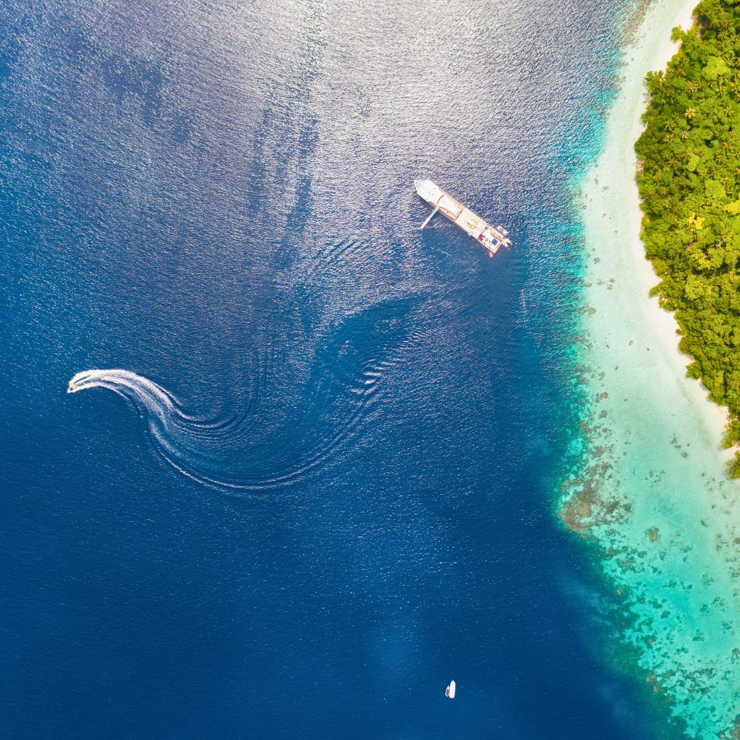 Aerial view of a turquoise sea with a sandy beach on one side and a small boat creating waves nearby. A larger ship is anchored in the distance. Lush greenery borders the shore.