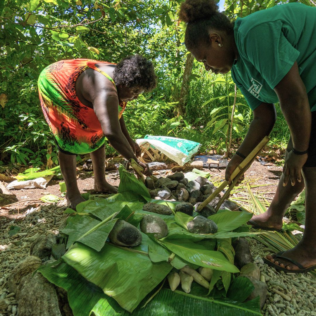 Two women preparing food using natural materials outdoors. One is arranging stones and leaves on the ground, while the other is adding more ingredients. Lush greenery surrounds them, creating a vibrant, tropical setting.