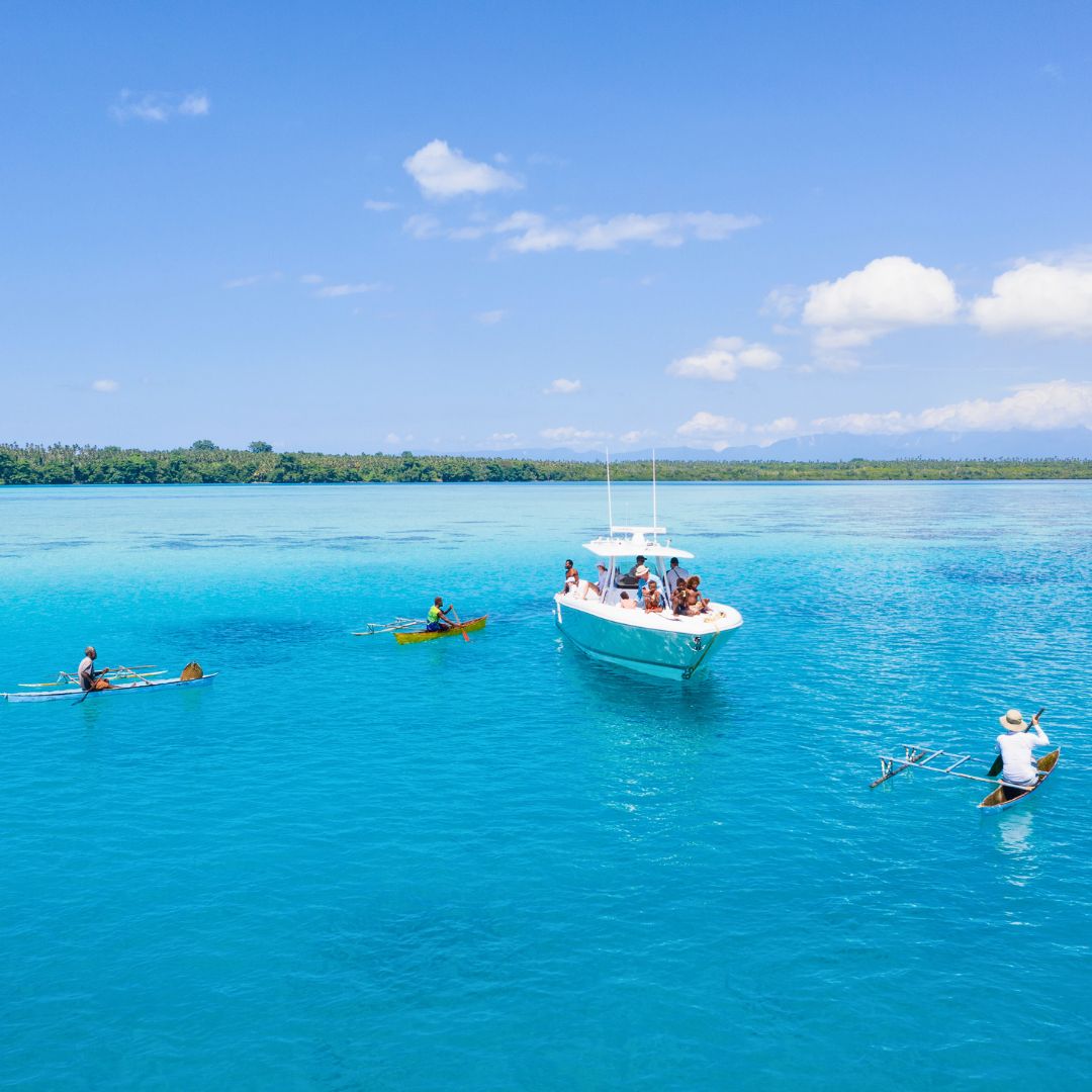 A boat with people on board floats in vibrant turquoise waters, surrounded by a lush green shoreline. Nearby, two individuals in small outrigger canoes paddle through the clear water under a bright blue sky with fluffy white clouds.