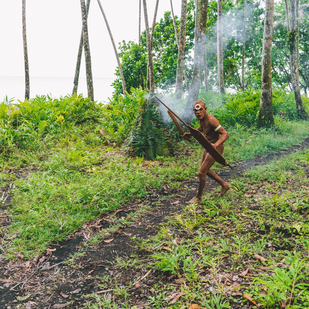 A person wearing traditional attire and a mask, holding a wooden spear, stands in a lush, tropical forest with smoke rising around them. They are surrounded by tall trees and greenery, with a view of the water in the background.