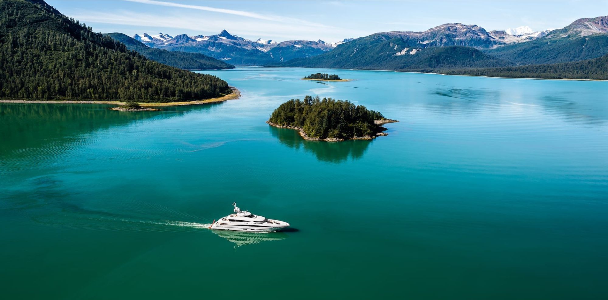 A white yacht cruises through calm turquoise water toward a forested island with snow-capped mountains in the distance.