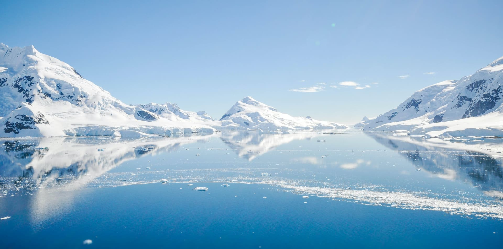 Snow-covered mountains reflect perfectly in the calm, mirror-like blue water of an icy polar bay under a clear sky.