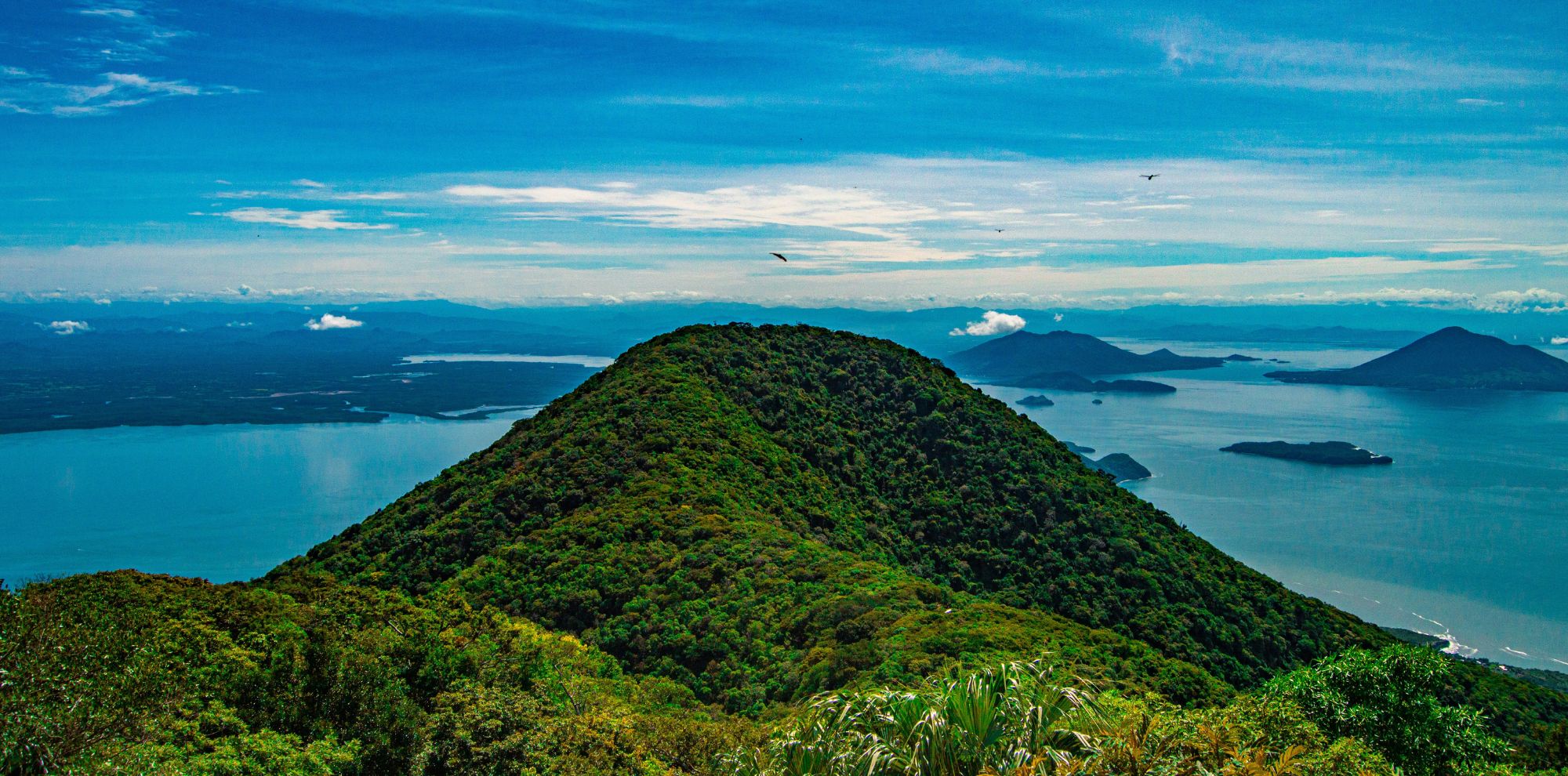 Lush green mountain peak overlooking a vast blue bay dotted with islands under a bright, clear sky.