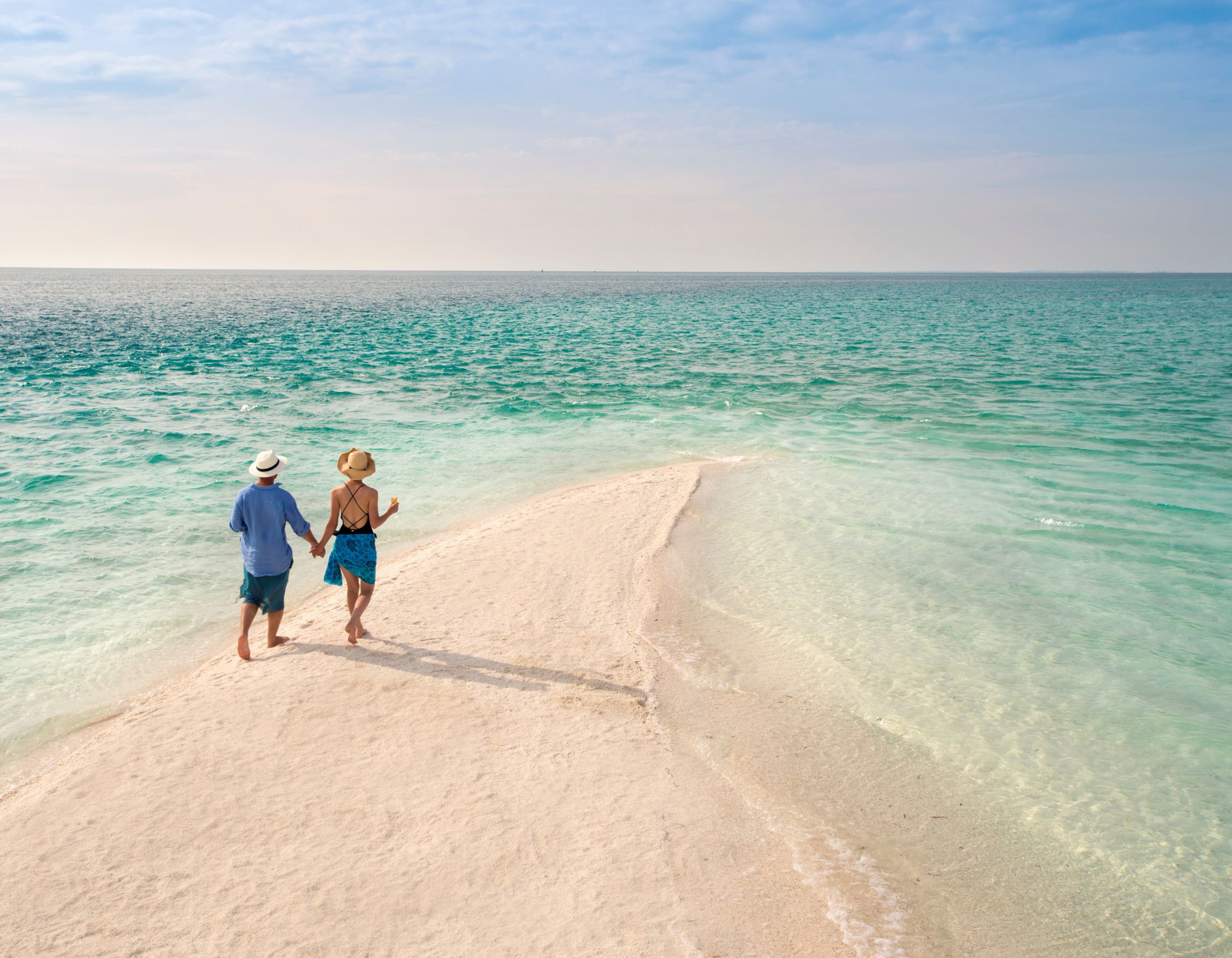 A couple holding hands while walking along a narrow, pristine white sandbar surrounded by turquoise tropical waters under a clear blue sky.