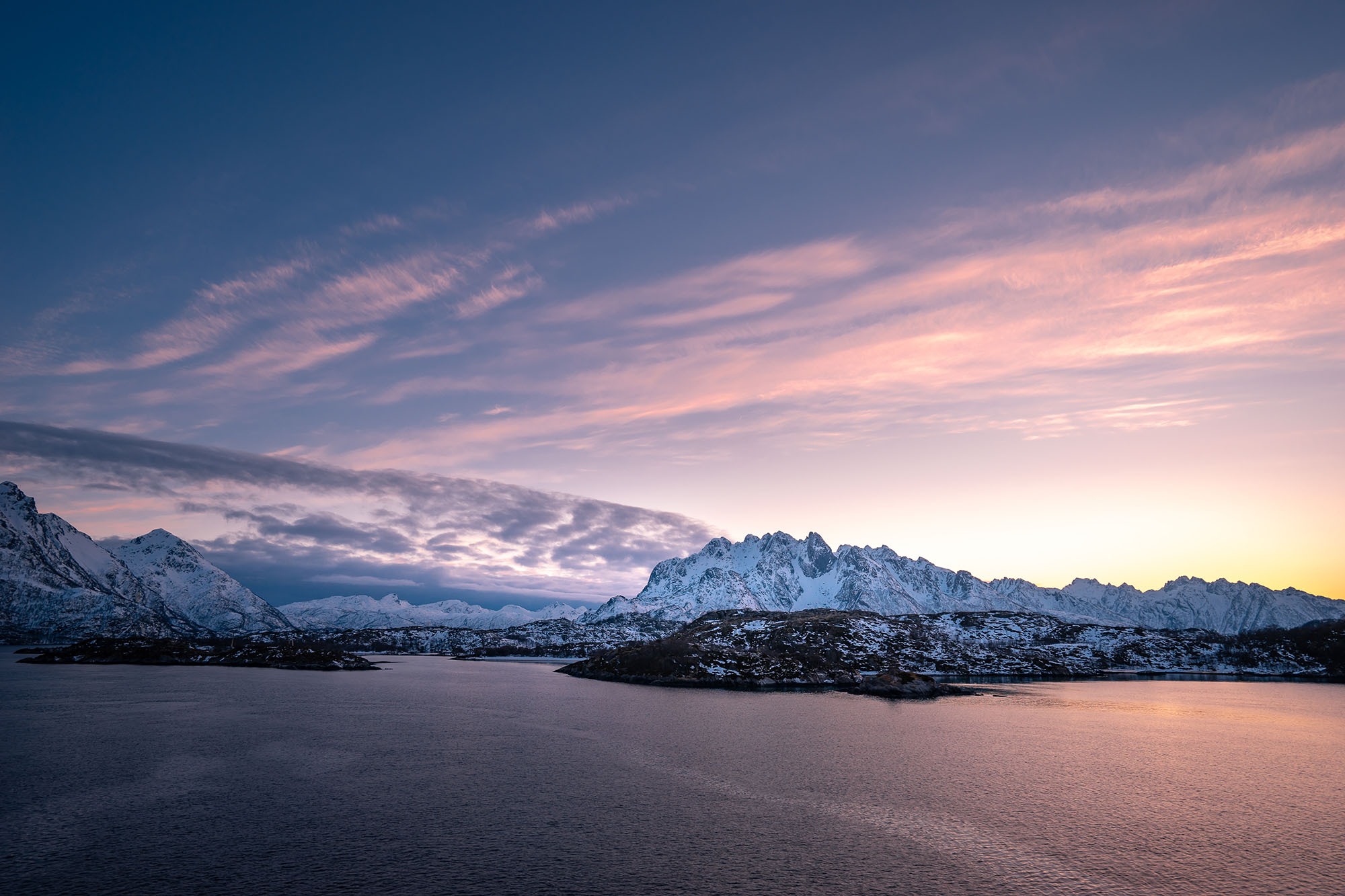 Panoramic view of a calm bay at dusk with snow-capped mountains under a sky filled with pink and purple clouds.