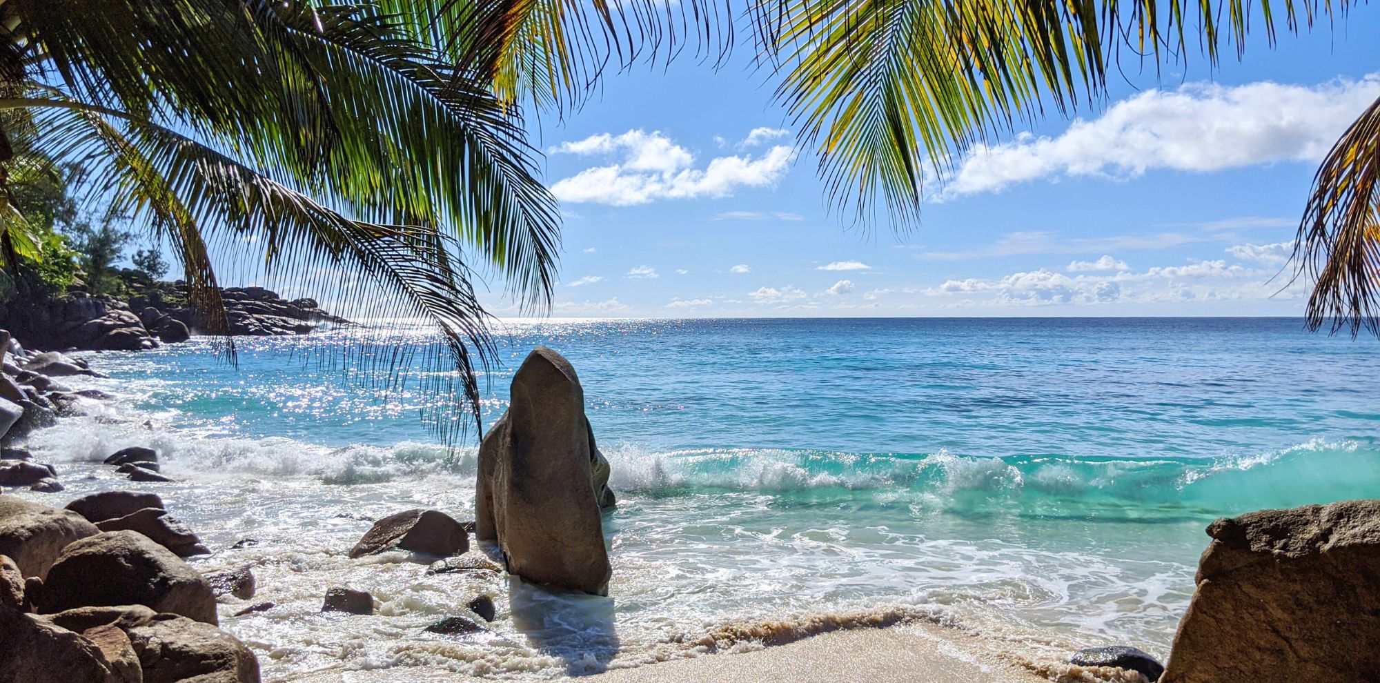 A scenic tropical beach view framed by palm fronds, featuring turquoise waves crashing against smooth granite boulders on a sun-drenched shore under a blue sky with scattered clouds.