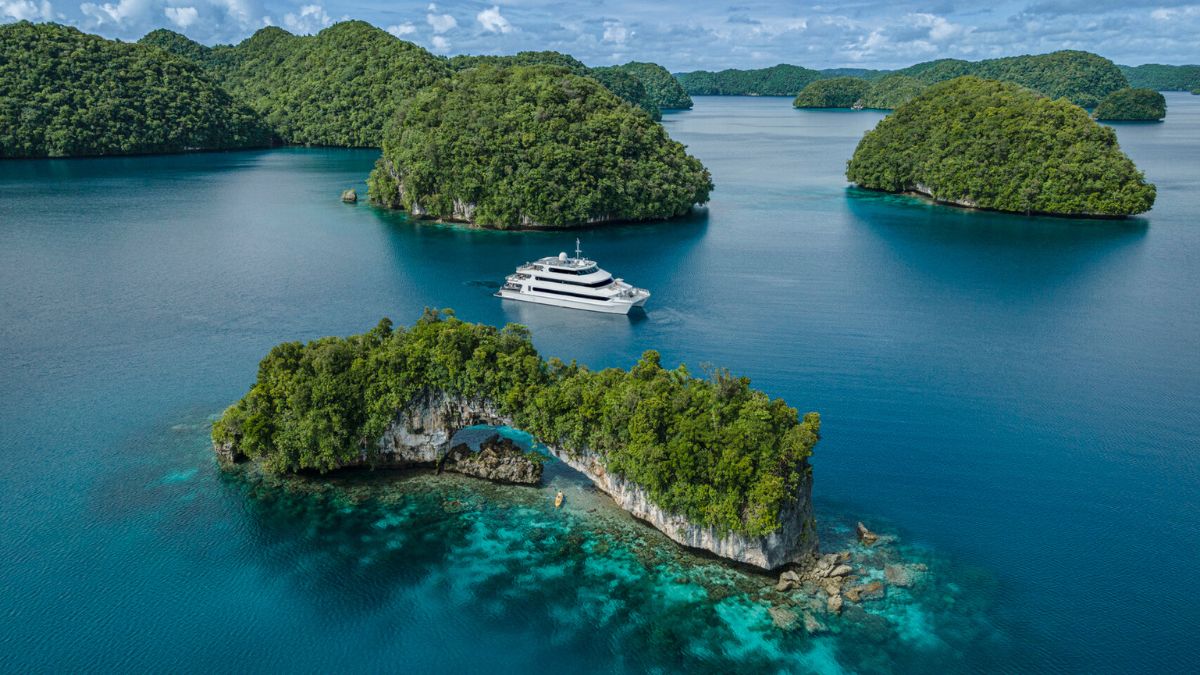 Aerial view of a white yacht cruising past lush green limestone islands in a clear blue tropical sea.