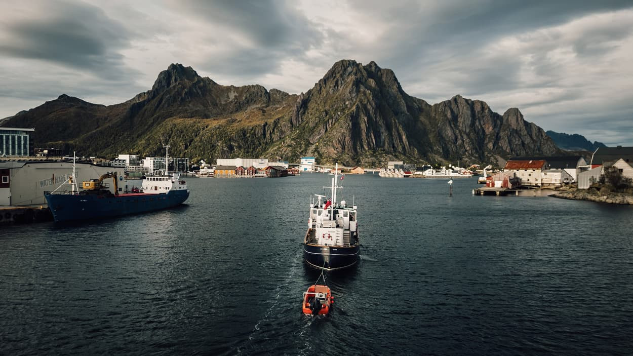 Small motor ship towing an orange lifeboat through a narrow fjord toward a coastal village at the foot of mountains.