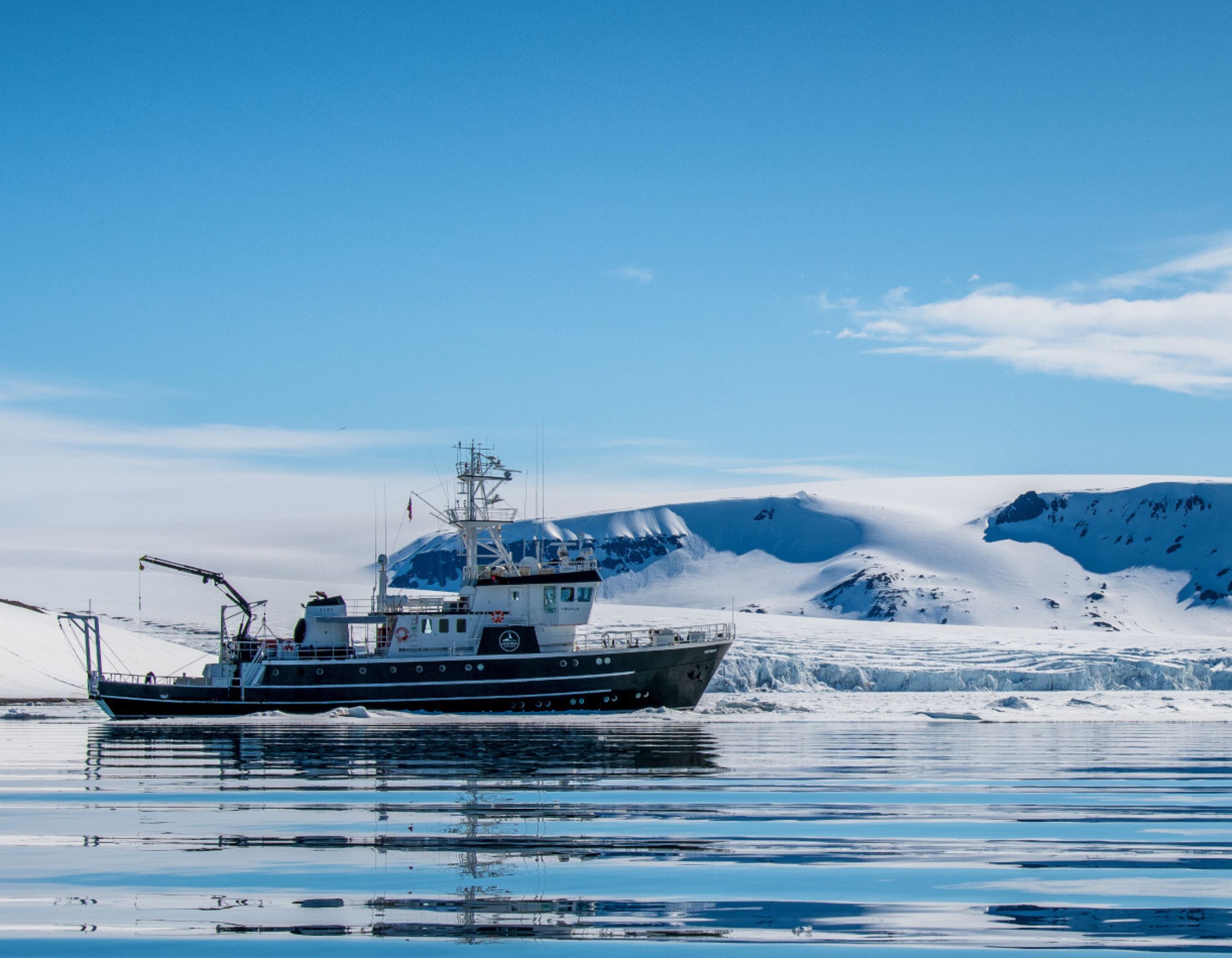 A black and white expedition ship sailing through icy arctic waters with glaciers and snow-covered hills behind.