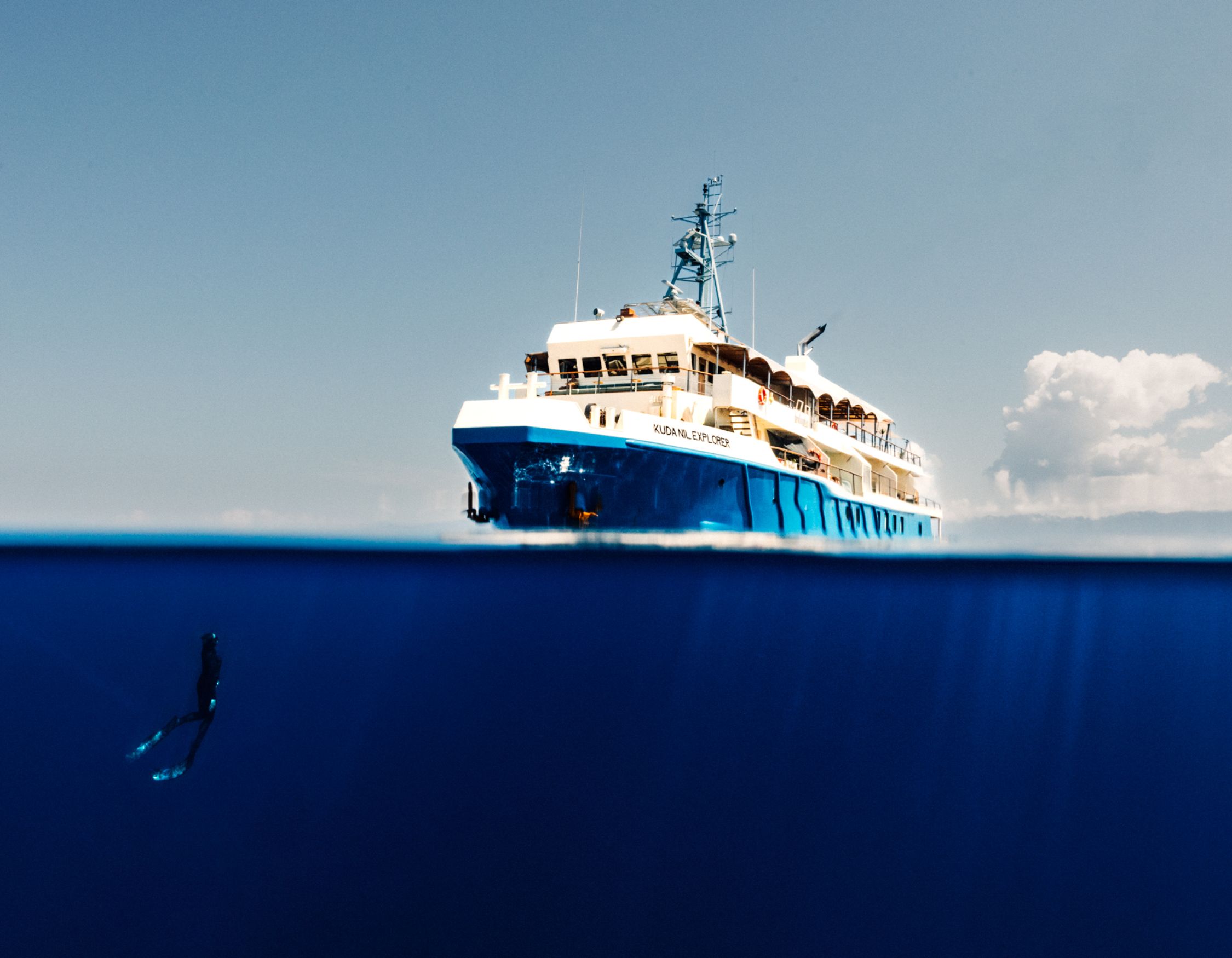 Split-level shot showing the motor yacht KUDANIL EXPLORER on the surface and a free-diver swimming in deep blue water below.