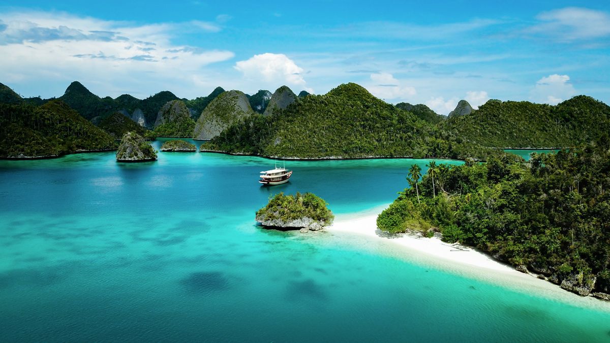 Aerial view of a wooden boat anchored in a turquoise lagoon surrounded by lush, green limestone hills and a white sand beach.