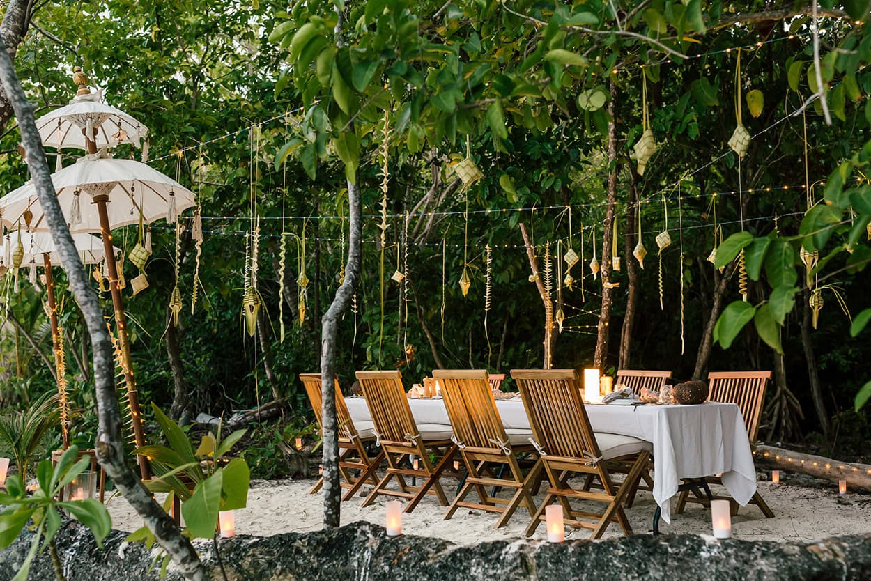 A long dining table with white linens and wooden chairs set on a beach under trees decorated with hanging ornaments and string lights.