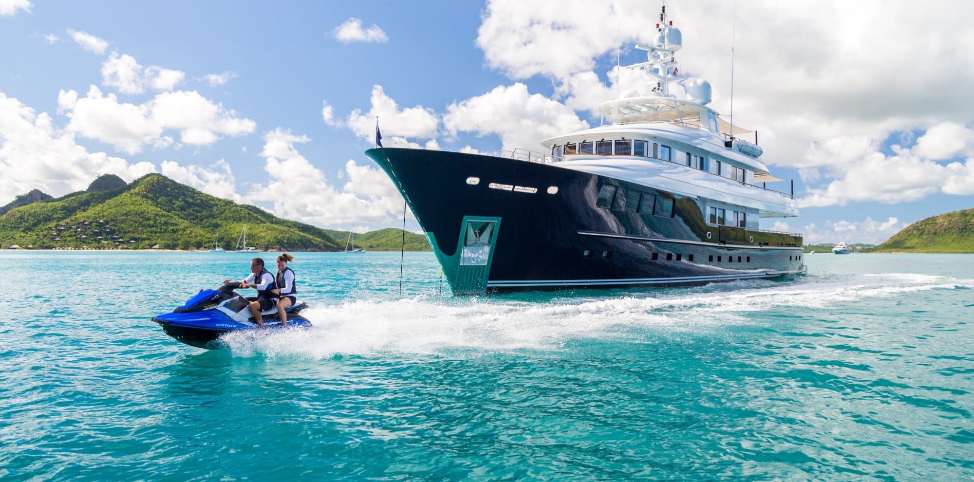 Two people on a blue jet ski speed past a large luxury yacht in clear turquoise water with green hills in the background.
