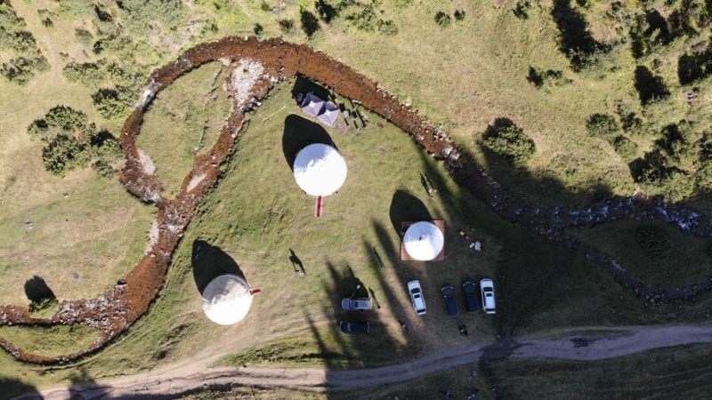 Aerial view of a natural landscape featuring three white yurt-like structures arranged on a grassy area. A winding stream flows nearby, with a few parked vehicles and people in the surroundings. Tall trees border the scene, creating a tranquil setting.