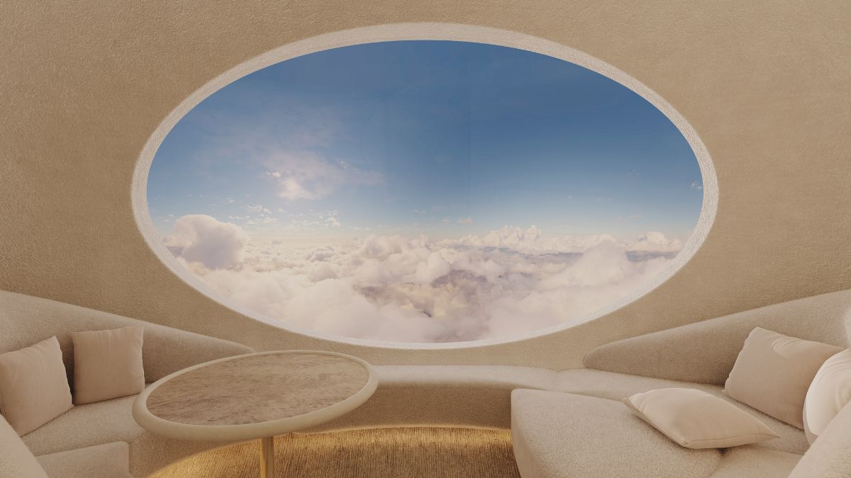 A cozy, modern interior with curved seating and a small table, featuring a large oval window that offers a panoramic view of fluffy clouds against a bright sky.