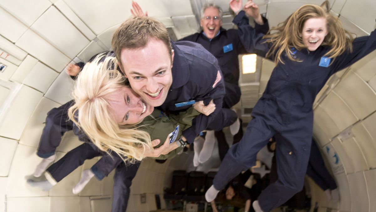 A group of four people in flight suits joyfully floating in a zero-gravity environment, with two individuals playfully embracing and the others laughing in the background.