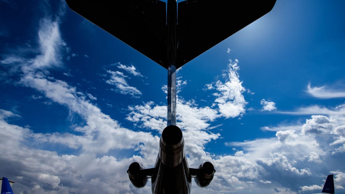 A jet's tail fin and engines are silhouetted against a bright blue sky filled with white clouds. The sun shines in the background, highlighting the contours of the aircraft.