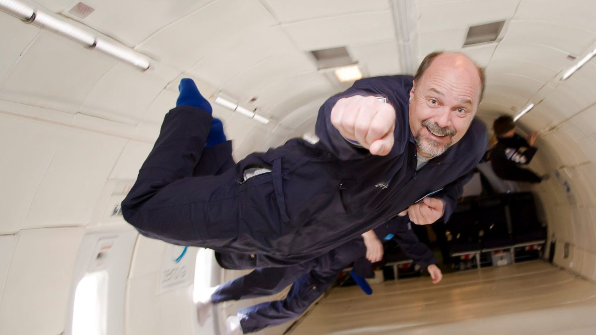 A man in a blue jumpsuit is floating inside an aircraft, smiling and making a fist as if celebrating. The interior shows the aircraft’s walls and several other people in the background, all experiencing weightlessness.