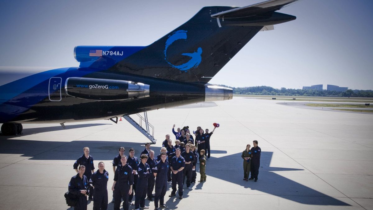 A group of people in blue uniforms stands in front of a large aircraft on a tarmac, smiling and waving. The aircraft has a blue logo and is marked with the registration number N794AJ. In the background, there are trees and buildings.