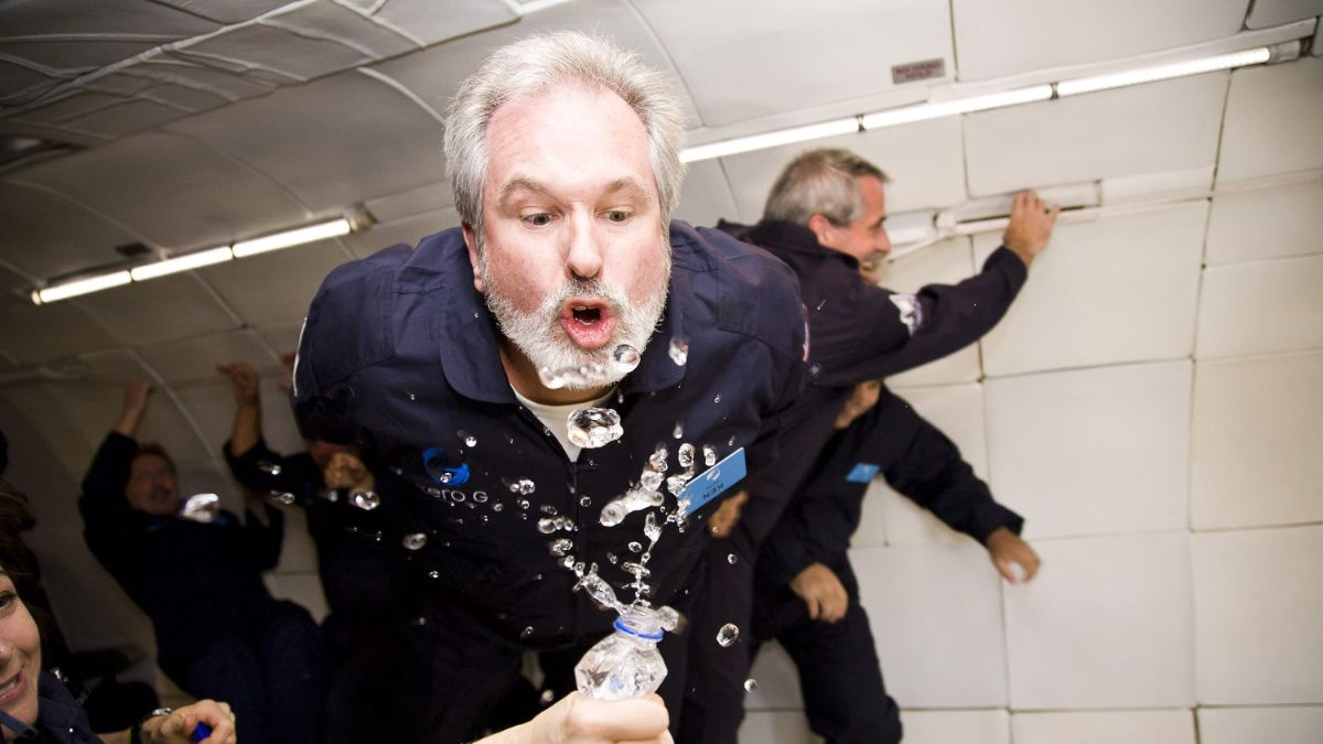 A man, wearing a blue flight suit, is in a zero-gravity setting, blowing bubbles from a water bottle. Several other people can be seen in the background, also enjoying the weightlessness.
